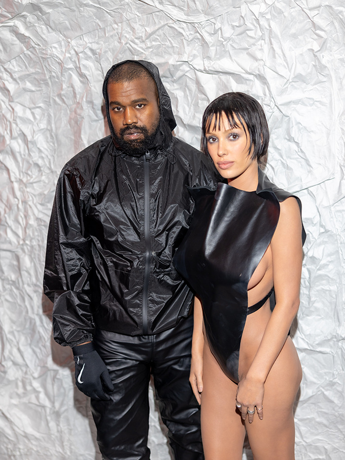 Kanye West and Bianca Censori poses in stylish black outfits against a textured backdrop.