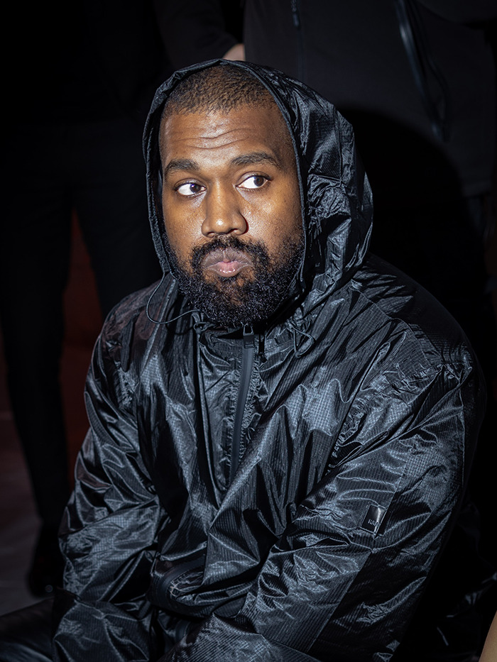 Kanye West in a black jacket sits indoors, appearing thoughtful, under low lighting.