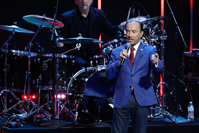 Lee Greenwood singing on stage with drummer in the background, wearing a blue blazer and red tie.