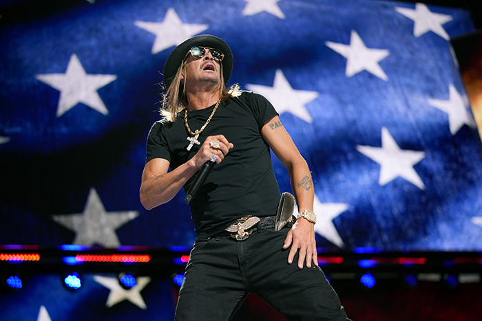 Kid Rock performing on stage, wearing a black outfit and hat, with an American flag backdrop.