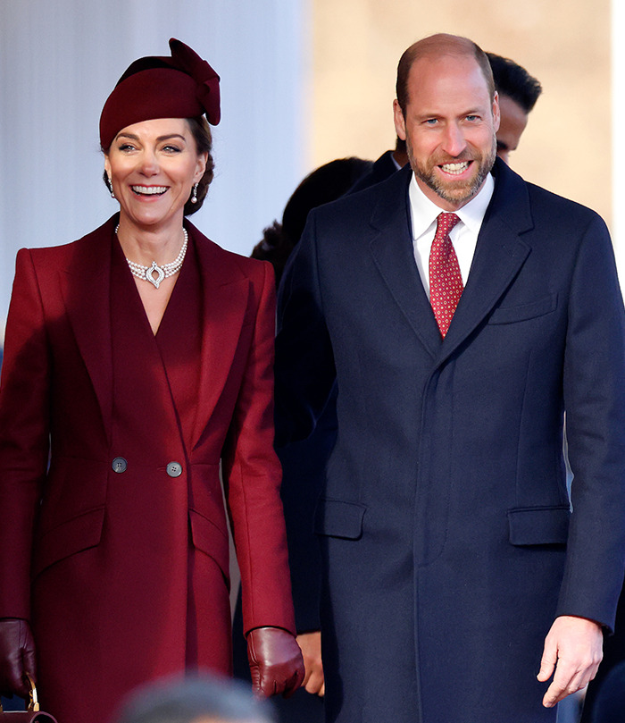 Prince William smiling in a suit beside Kate Middleton in a burgundy coat and hat on a sunny day.