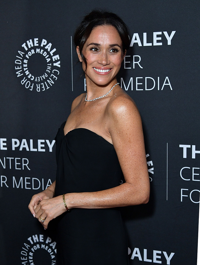 Meghan Markle in black dress at media event, smiling in front of a branded backdrop. Meghan Markle in black dress at media event, smiling in front of a branded backdrop.