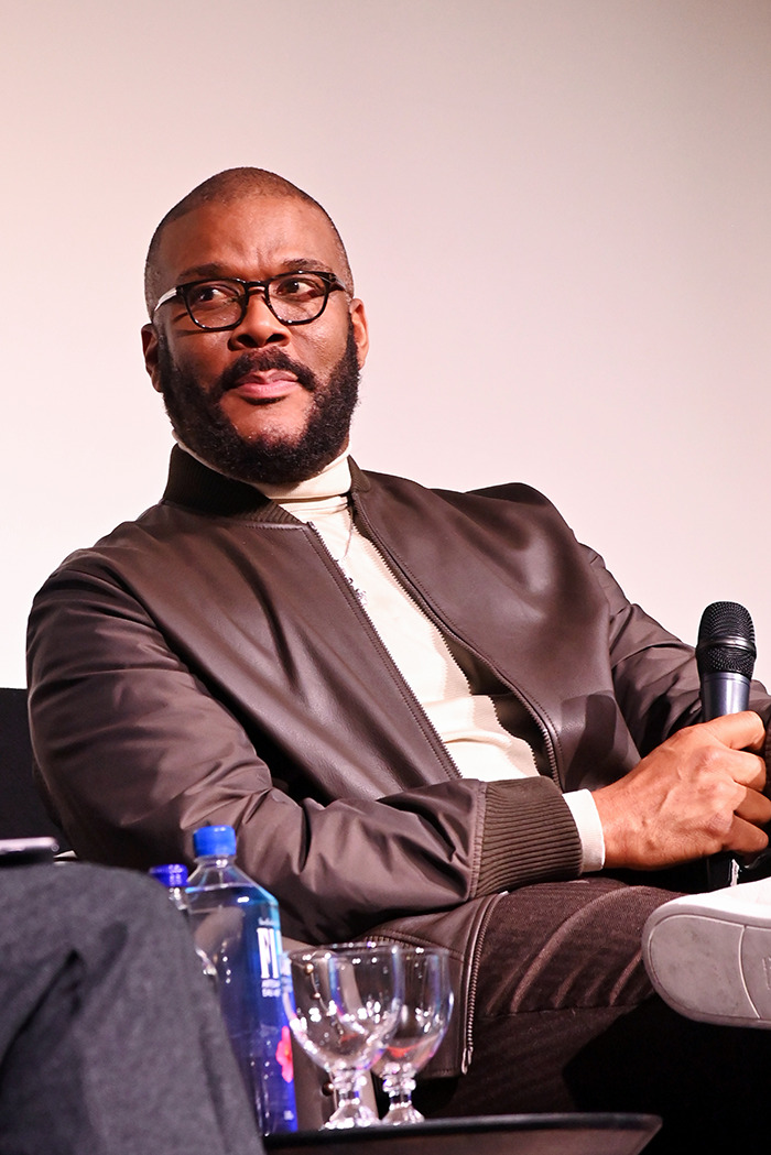 Tyler Perry speaking at an event, holding a microphone, wearing glasses and a brown jacket.