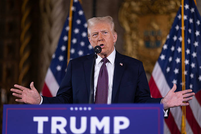 Donald Trump speaking at a podium with "TRUMP" sign in front, against a backdrop of American flags.