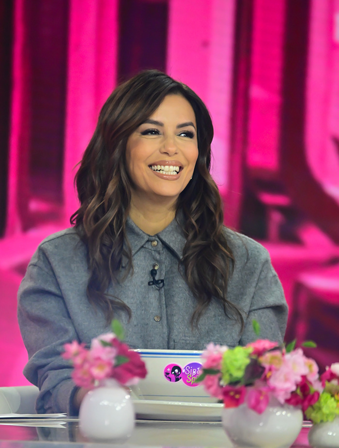 Eva Longoria smiling on a talk show set with pink background and floral decorations.
