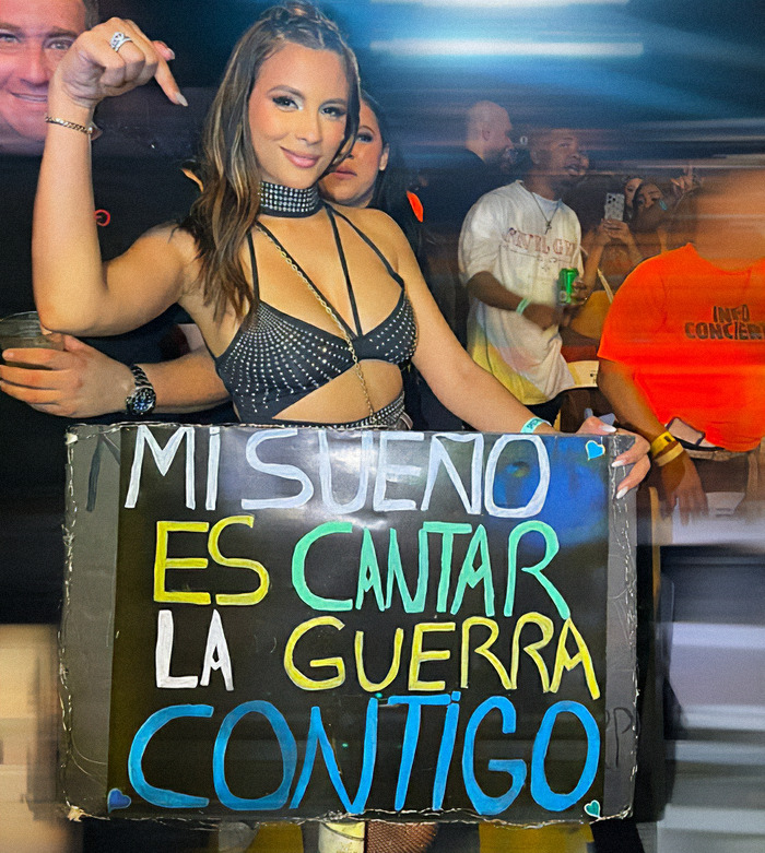Influencer at concert holding a sign, surrounded by a lively crowd, with colorful lights and energy.