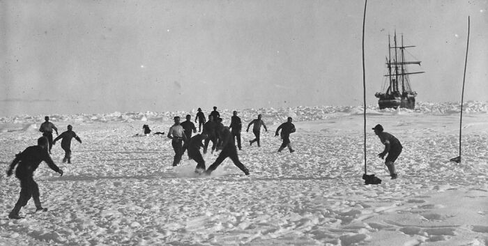 People playing soccer on snow with a ship in the background during the Imperial Trans-Antarctic Expedition.