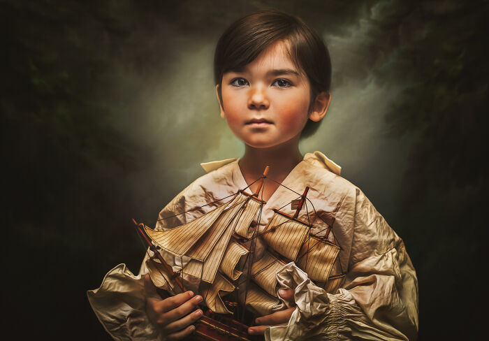 Creative photo of a child holding a model ship, wearing a vintage-style shirt against a moody background.