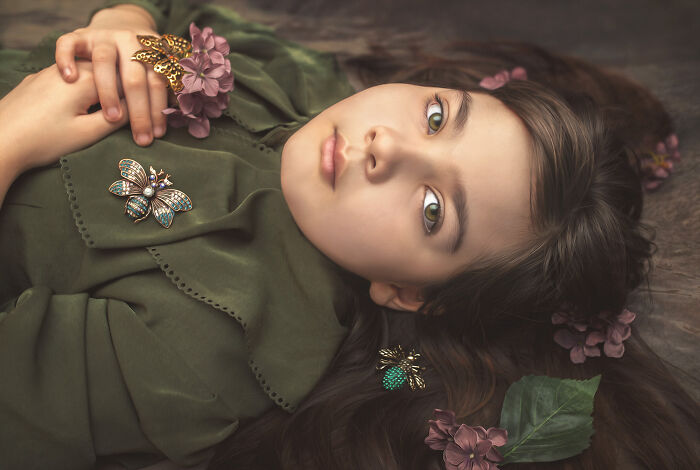 Child in green dress lying down, adorned with flowers and butterfly accessories, showcasing creative photography.