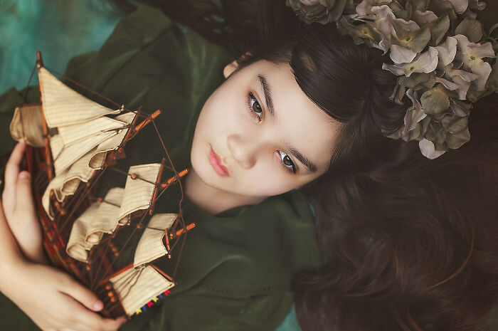 Child holding a model ship, lying down with flowers in hair, in creative photography setting.