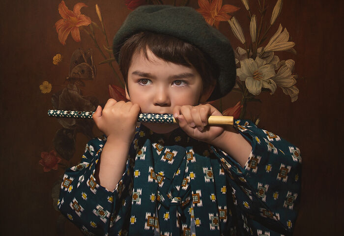 Child in a patterned outfit with a beret, holding a flute, surrounded by floral wallpaper. Creative kids photography.