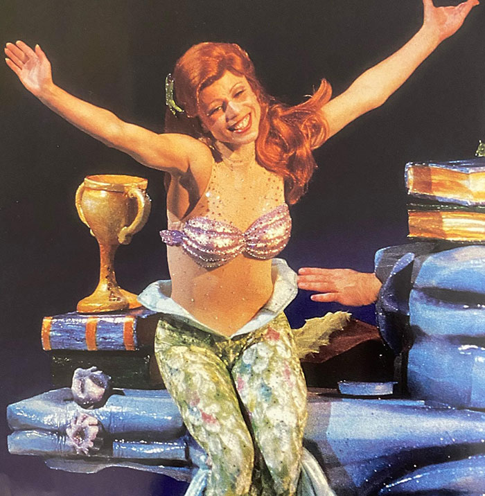 Disney On Ice actress performing as a mermaid with arms raised, surrounded by colorful stage props.