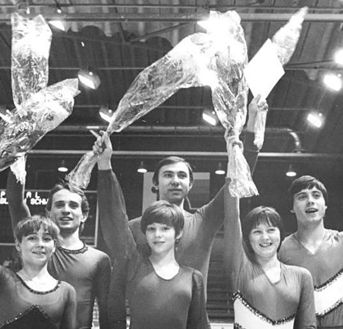 Performers in 'Disney On Ice' costumes celebrating with bouquets, indoors, under a lit ceiling.