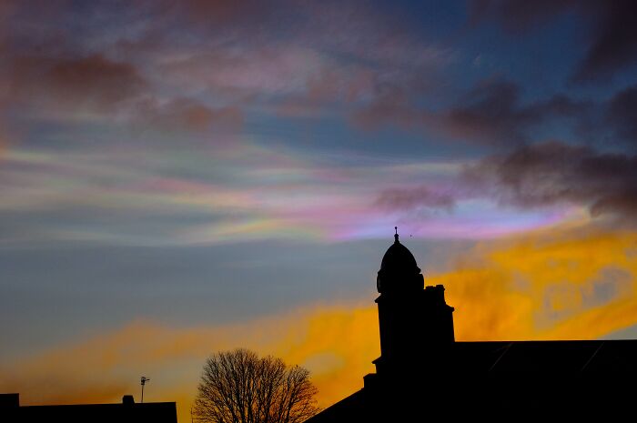 Colorful iridescent clouds in the sky above a silhouette of buildings and trees, showcasing stunning natural phenomena.