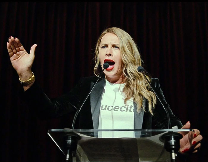 Transgender Oscar nominee speaking passionately at a podium with a raised hand.