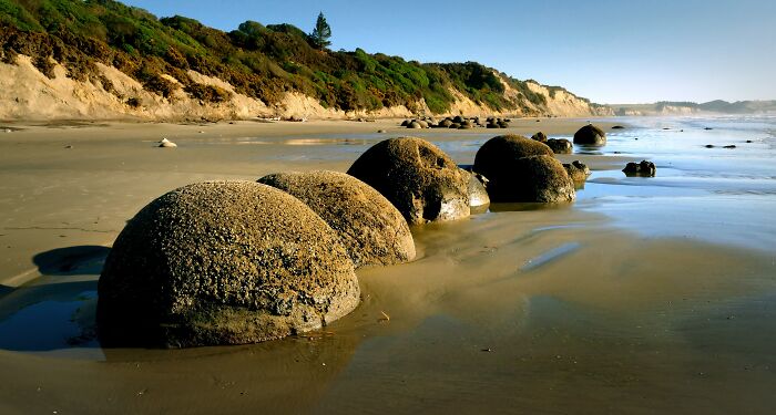 Natural phenomena: Spherical boulders on a sandy beach under a clear sky with coastal vegetation in the background.