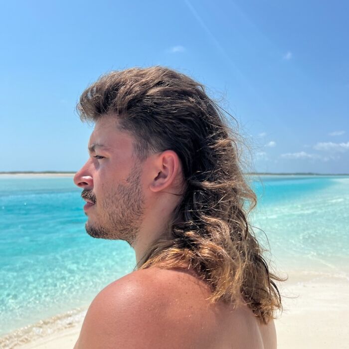 Person with mullet hairstyle, standing by the beach with clear blue water under a sunny sky.