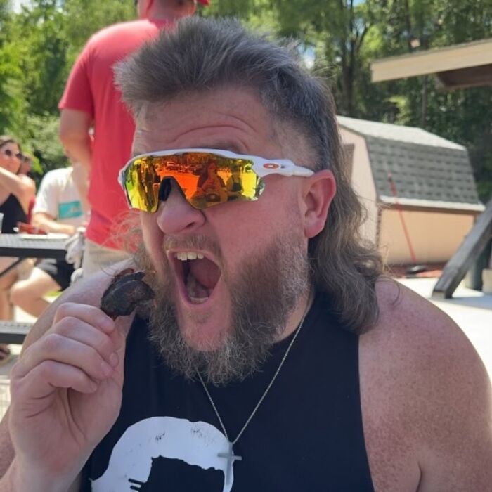 Man with mullet hairstyle wearing sunglasses, outdoors, eating.