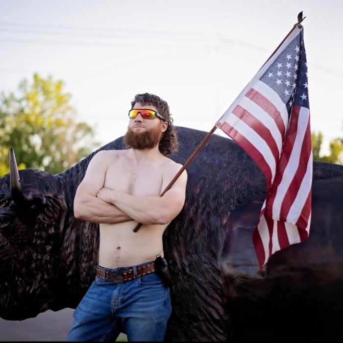 Man showcasing a mullet hairstyle, wearing sunglasses, holding an American flag, standing confidently outdoors.