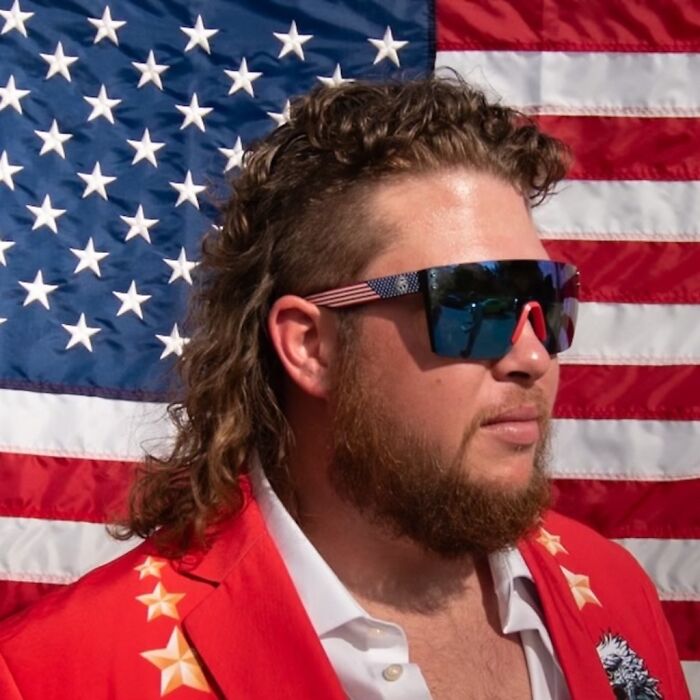 Person with mullet hairstyle wearing sunglasses and a red jacket, standing in front of an American flag.