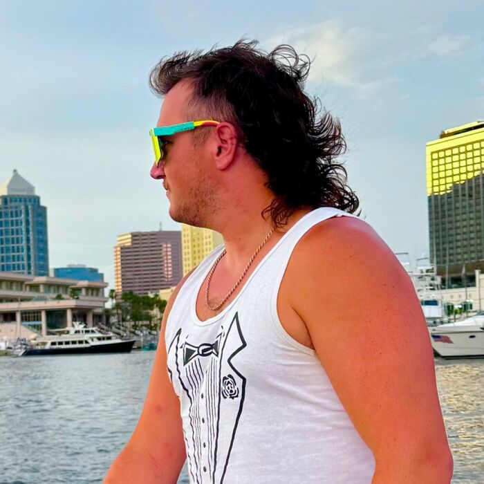 Man with mullet hairstyle on a waterfront, wearing sunglasses and a tuxedo-print tank top, showcasing a stylish 2024 hairdo.