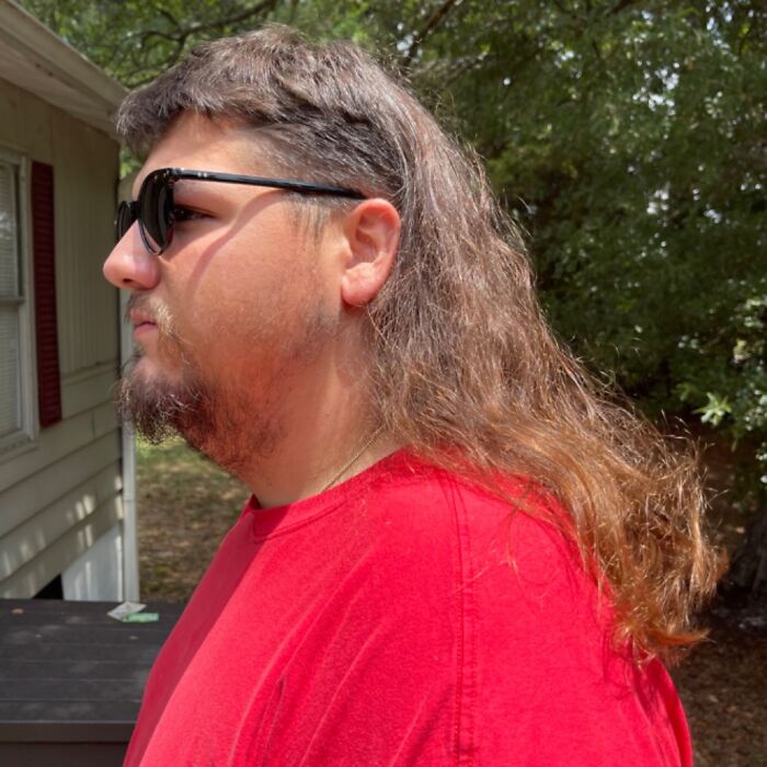 Person with a mullet hairstyle wearing sunglasses and a red shirt outdoors.