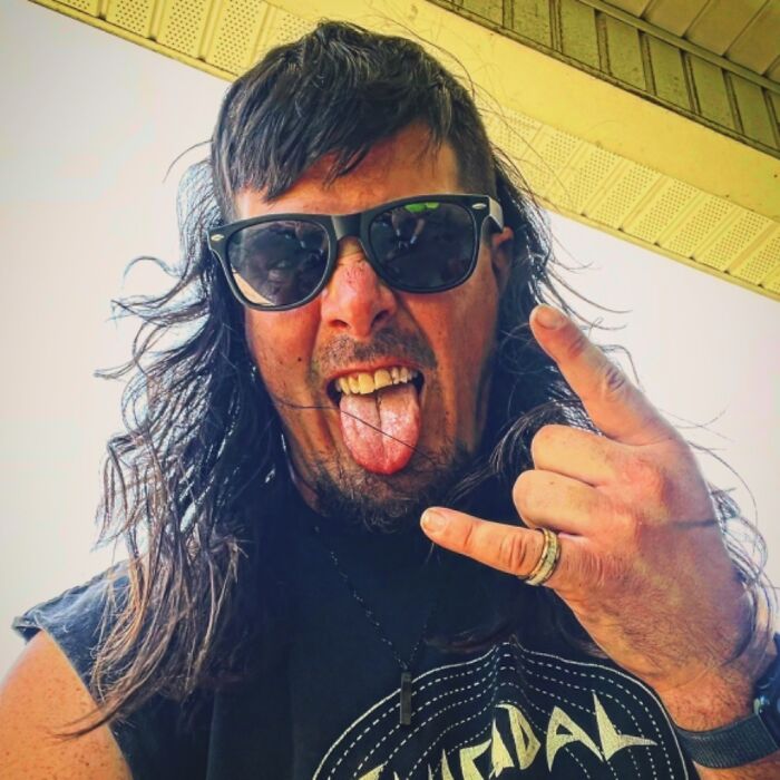 Person with a mullet hairstyle wearing sunglasses, making a rock hand gesture, and sticking out their tongue.