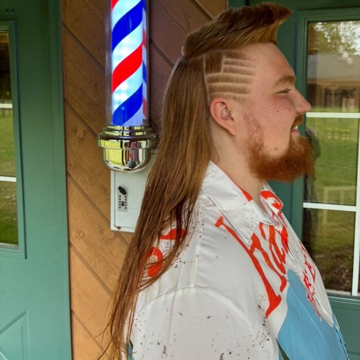Man with a stylish mullet standing outside a barber shop, showcasing long hair in the back and shaved designs on the side.