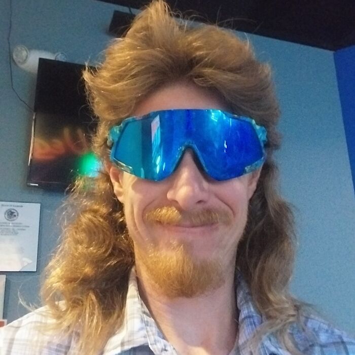 Man with a mullet hairstyle wearing reflective sunglasses and a checkered shirt, smiling indoors.