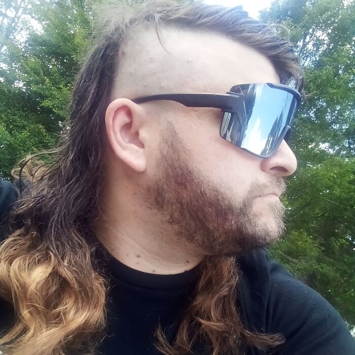 Man with mullet hairstyle wearing reflective sunglasses, outdoor setting.