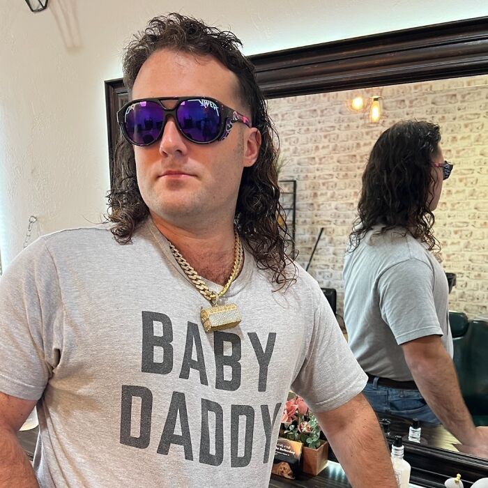 Person with mullet hairstyle, wearing sunglasses and a "BABY DADDY" shirt, posing in a stylish setting.