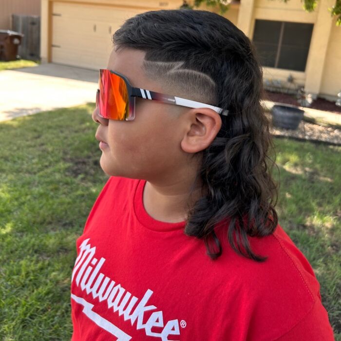 Child wearing sunglasses and a red shirt with a stylish 2024 mullet haircut outdoors.