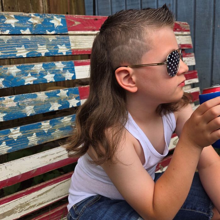 A child with a mullet hairstyle, wearing star-spangled sunglasses and holding a drink, sits on a patriotic bench.
