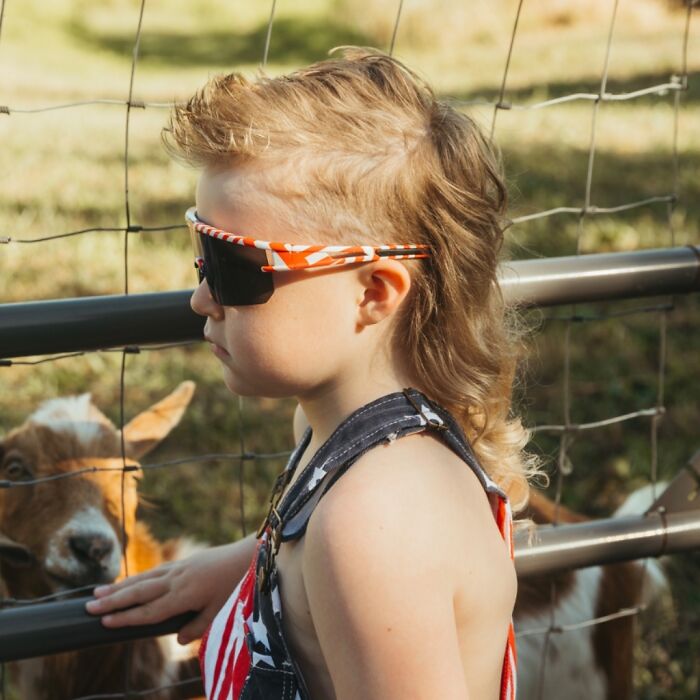 Child with a stylish mullet hairstyle, wearing sunglasses, standing next to a goat by a fence.