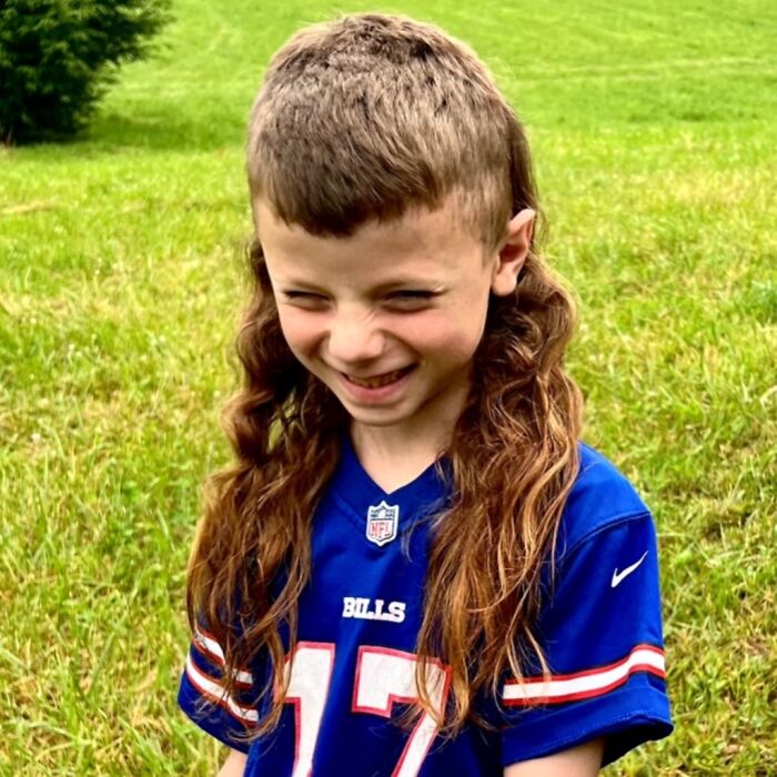 Child with a mullet hairstyle smiles in a football jersey against a grassy backdrop.
