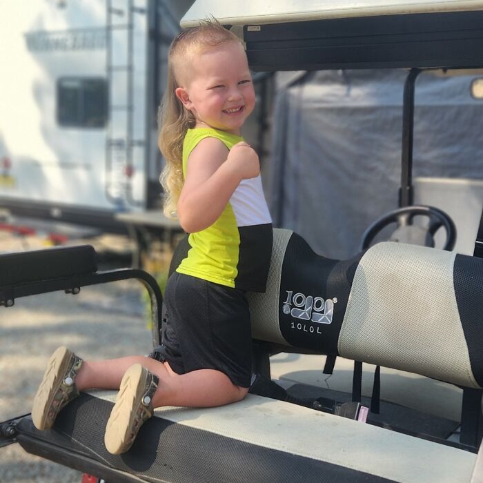 Child with a stylish mullet hairstyle smiling while kneeling on a golf cart seat, wearing a bright yellow shirt.