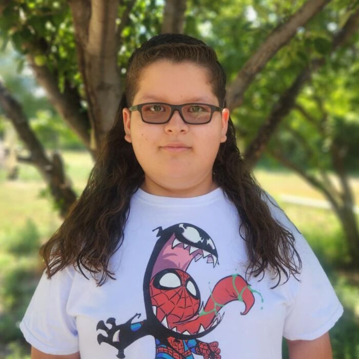 A young person with glasses and long hair in a mullet style, wearing a Spiderman t-shirt, standing outdoors.