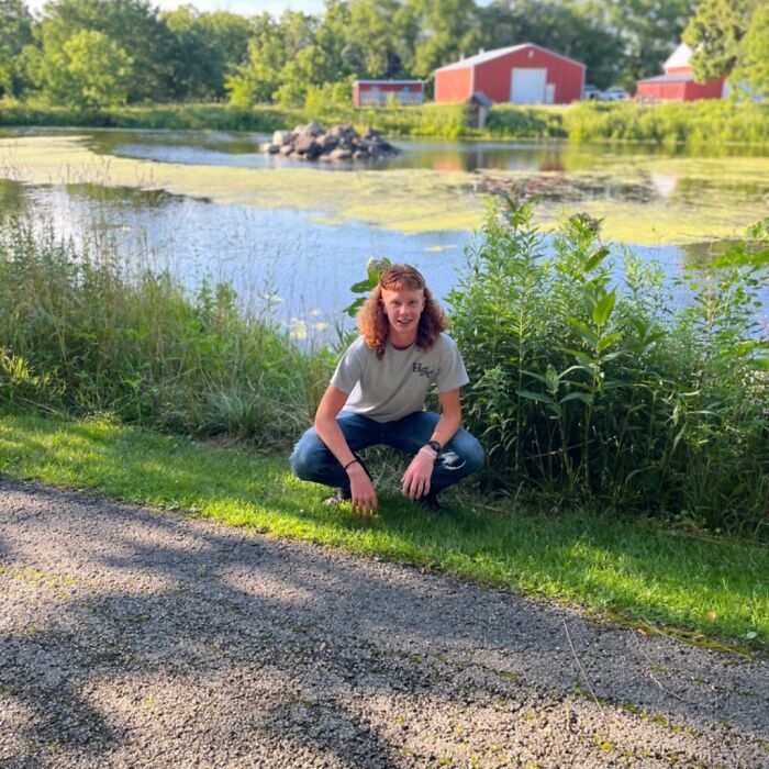 Individual with a mullet hairstyle crouching by a picturesque pond, surrounded by greenery and red barns in the background.