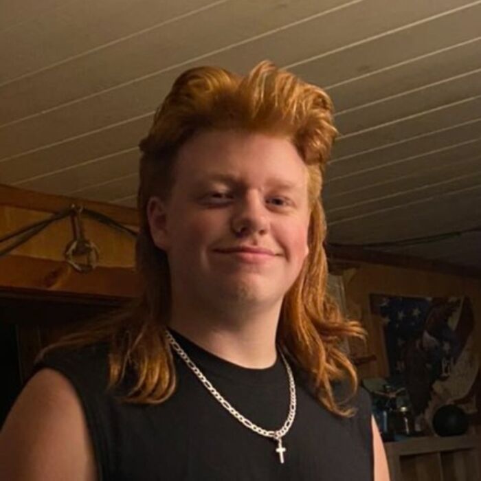 Person with a stylish mullet hairstyle, wearing a black sleeveless shirt and silver chain, inside a rustic room.
