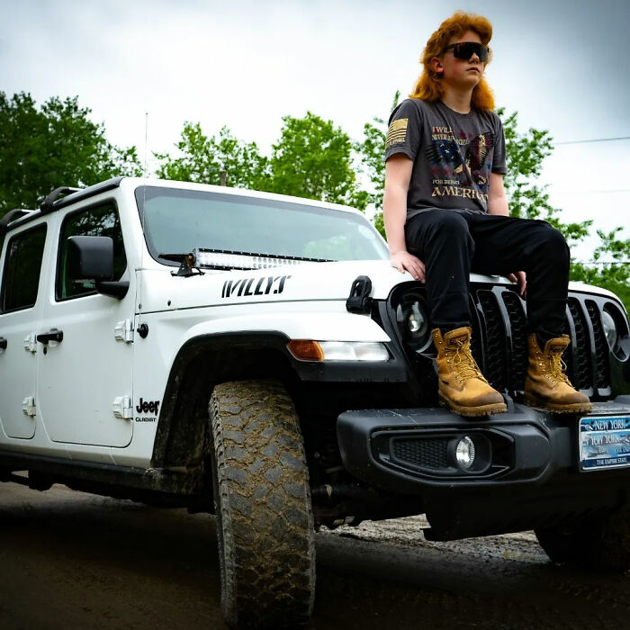 Young person with mullet hairstyle sitting on a Jeep, wearing sunglasses and boots, outdoors.