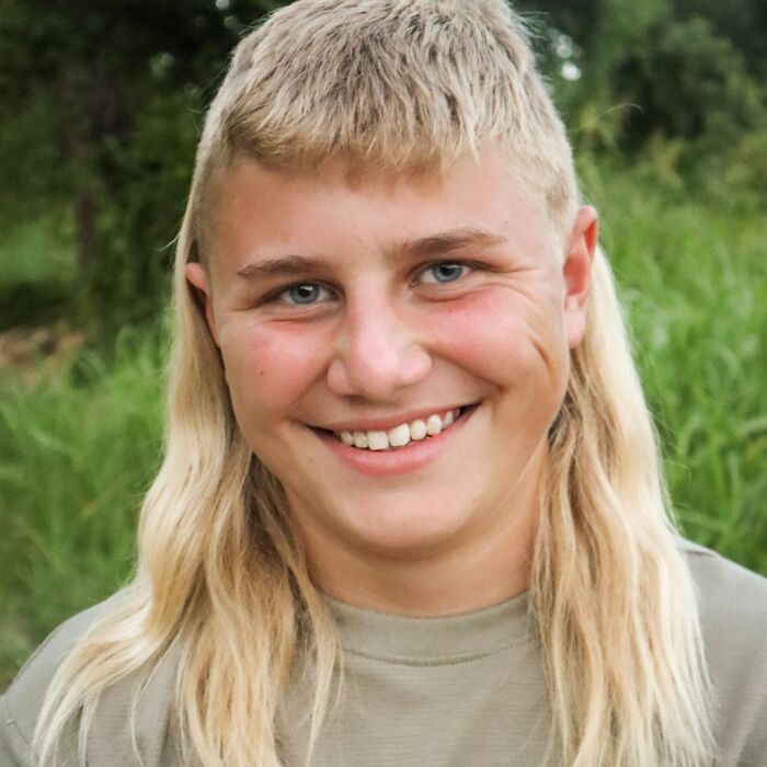 Smiling person with a mullet hairstyle in a green outdoor setting.