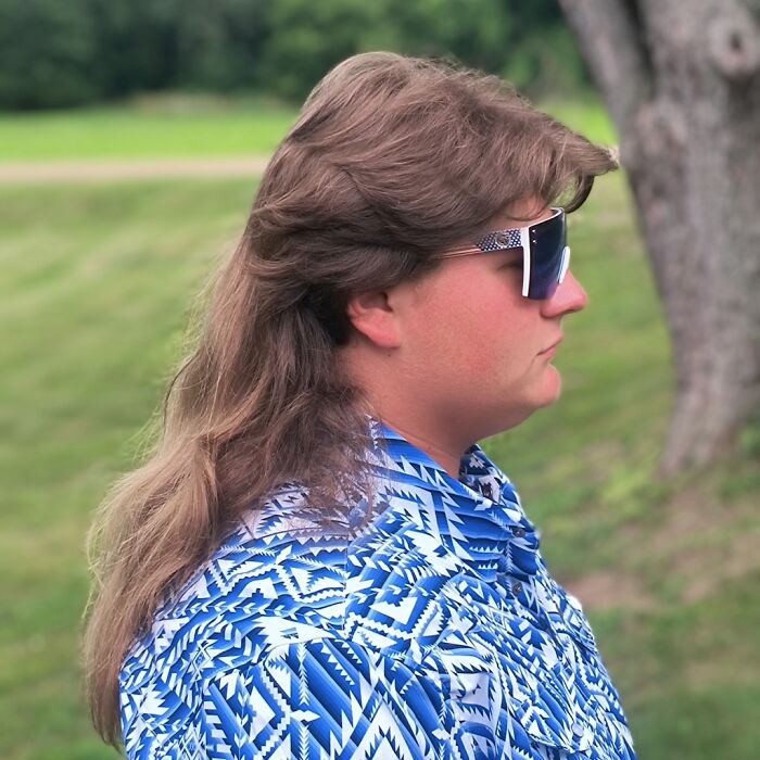 Person with mullet hairstyle, wearing patterned shirt and sunglasses, outdoors.