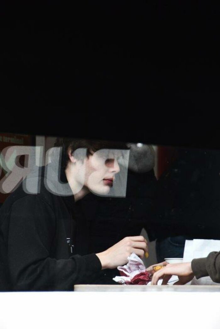 Person seen through a window, holding food, with a logo partially visible, capturing everyday moments through Nadia Eeckhout’s lens.