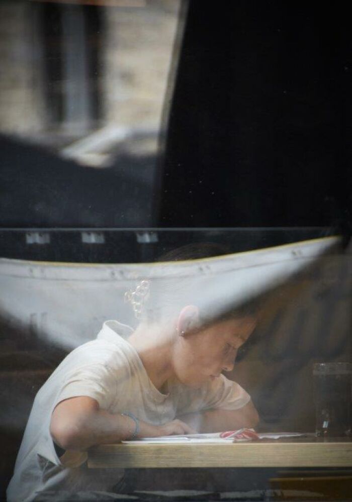 Person reading at a table, captured through glass, showcasing everyday moments by Nadia Eeckhout’s lens.