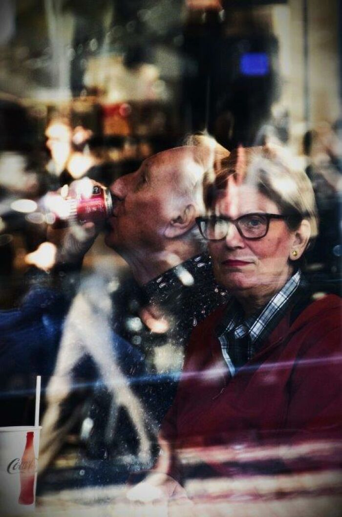 Elderly couple through a cafe window, with a man sipping a drink, capturing everyday moments through Nadia Eeckhout's lens.