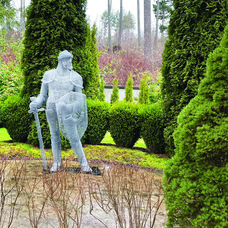 Chicken wire sculpture of a knight holding a sword and shield, set in a garden with lush greenery.