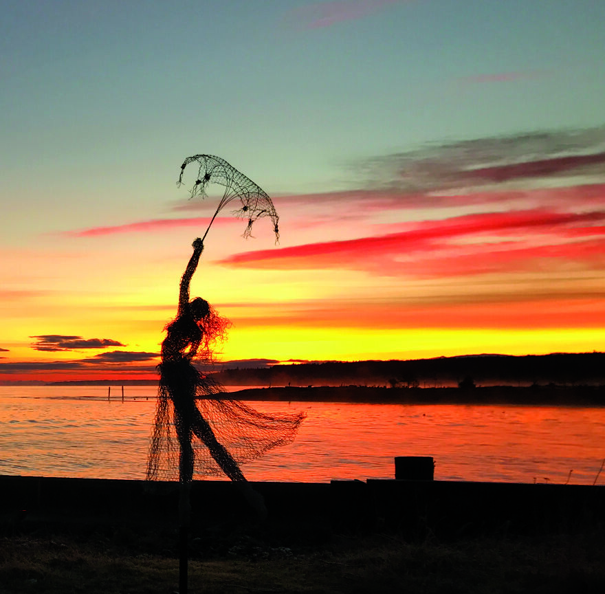 Chicken wire sculpture depicting a dancer with an umbrella against a vibrant sunset by the water.
