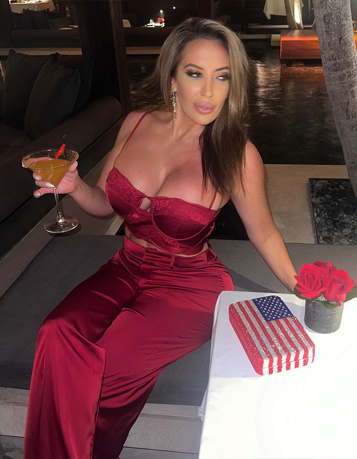 Adult star in red outfit seated at a table, holding a cocktail, with patriotic decoration and roses nearby.