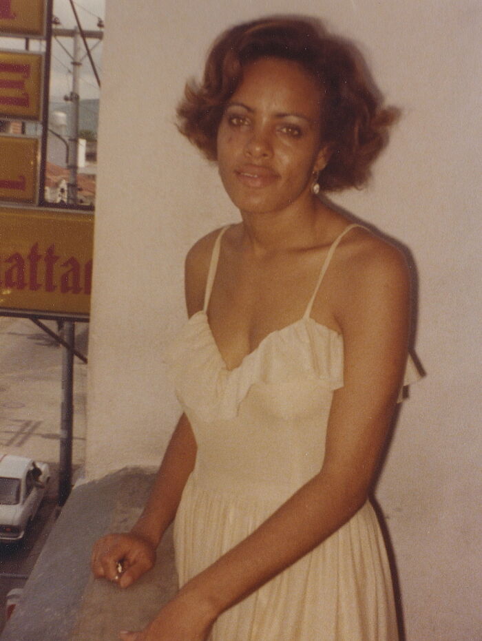 Woman in 80s fashion wearing a ruffled dress, standing on a balcony with a city street and signage in the background.
