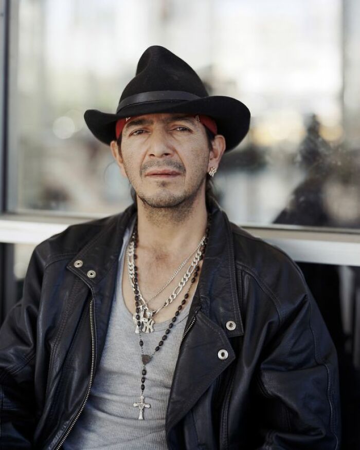 Man wearing a black hat and leather jacket sitting at an America bus stop in a candid street portrait.